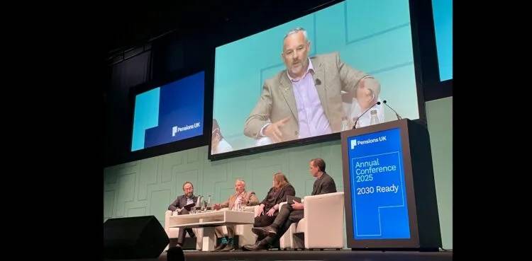 A panel discussion taking place on a stage at a conference. Four individuals are seated in white armchairs around a small table with water bottles and glasses. Behind them is a large screen displaying a close-up of one of the speakers. To the right of the stage is a tall blue sign that reads: “Pensions UK Annual Conference 2025 2030 Ready.”