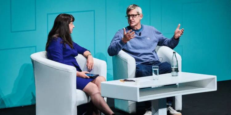 Two individuals seated in white armchairs on a stage with a teal backdrop, engaged in a discussion. A small white table between them holds a glass of water, a bottle of water, and a notepad. One person is wearing a blue outfit and holding a tablet, while the other is dressed in a light blue sweater and gesturing with both hands.