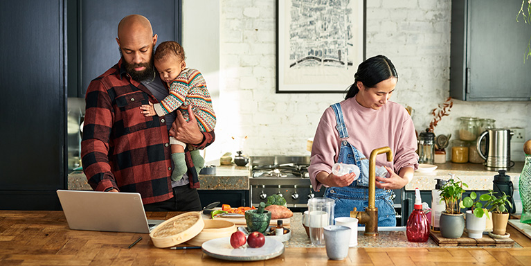 Man holding baby in kitchen using laptop